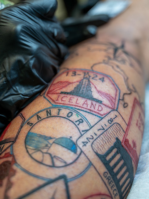 A colorful travel-themed tattoo on the leg, featuring detailed stamps from Iceland, Santorini, and Greece, symbolizing personal journeys and exploration.A colorful travel-themed tattoo on the leg, featuring detailed stamps from Iceland, Santorini, and Greece, symbolizing personal journeys and exploration.
